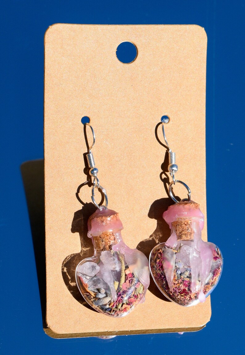 May include: A pair of heart-shaped glass bottle earrings with cork stoppers. The bottles are filled with pink and white crystals, dried flowers, and herbs. The earrings are hanging from silver hooks.