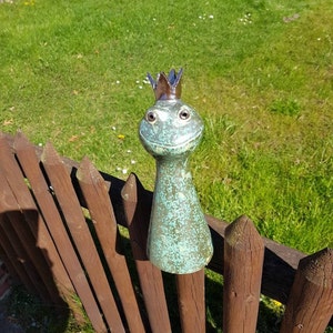 Könnte beinhalten: Eine skurrile, türkisfarbene Froschstatue mit einer bronzenen Krone, die auf einem Holzzaun sitzt. Der Frosch hat große, ausdrucksstarke Augen und einen lächelnden Mund. Die Statue steht vor dem Hintergrund von grünem Gras und einem sonnigen Tag.