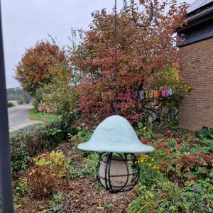 Könnte beinhalten: Ein blauer Vogel futterspender mit einem runden Käfig und einem hellblauen Dach, das an einem Baumzweig in einem Garten hängt.