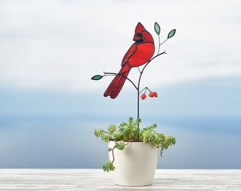 May include: A stained glass cardinal perched on a branch with red berries, mounted on a black metal stake. The cardinal is red with black accents and is positioned in front of a potted succulent plant.