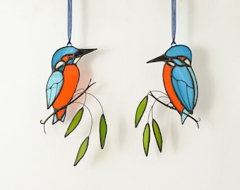 Könnte beinhalten: Zwei Buntspechtvögel aus Buntglas, einer mit blauen und orangen Flügeln und der andere mit blauen und grünen Flügeln, sitzen auf Ästen mit grünen Blättern.