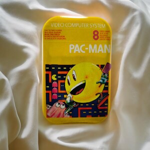 Könnte beinhalten: Eine rechteckige gelbe Blechdose mit einer Pac-Man-Grafik. Der Text lautet VIDEO COMPUTER SYSTEM, 8 VIDEO GAMES und PAC-MAN. Das Design zeigt eine gelbe Pac-Man-Figur, rosa Geister und ein Labyrinth-Muster auf einem dunklen Hintergrund.