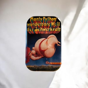 Könnte beinhalten: Eine rechteckige Metalldose mit dem Filmplakat von Monty Python's wunderbare Welt der Schwerkraft. Das Bild zeigt eine große, nackte Figur in einem roten Tanga, die vor einem dunkelblauen Hintergrund schwebt, mit gelbem Text oben.
