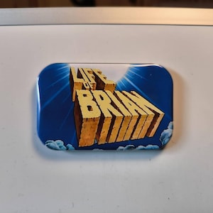 Könnte beinhalten: Ein rechteckiger Ansteckbutton mit blauem Hintergrund, weißen Wolken und Sonnenstrahlen. Der Schriftzug LIFE OF BRIAN ist in großen, dreidimensionalen, beigefarbenen Blockbuchstaben, die wie aus Stein gemeißelt wirken, mittig abgebildet.
