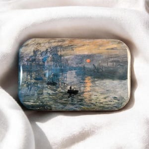 Könnte beinhalten: Eine rechteckige Anstecknadel mit einer Reproduktion von Claude Monets Gemälde Impression, Sonnenaufgang. Das Kunstwerk zeigt eine Hafenszene mit einem kleinen schwarzen Boot auf dem Wasser, Blau- und Grautönen sowie einer hellen orangefarbenen Sonne.