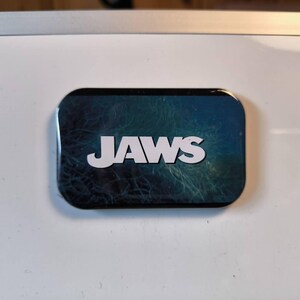Könnte beinhalten: Ein rechteckiger Magnet mit abgerundeten Ecken, der das Wort JAWS in fetten, weißen Großbuchstaben zeigt, zentriert auf einem dunklen, strukturierten blau-grünen Hintergrund, der an tiefes Meerwasser mit subtilen, wirbelnden Mustern erinnert.