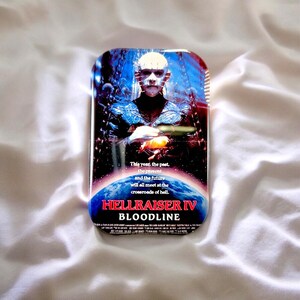 Könnte beinhalten: Eine rechteckige Metalldose mit dem Filmplakat von Hellraiser IV Bloodline. Das Design zeigt eine blasse Figur mit Nadeln im Kopf vor einem dunklen Hintergrund mit Ketten, einer leuchtend orangefarbenen Kugel und dem Text: This year, the past, the present and the future will all meet at the crossroads of hell. HELLRAISER IV BLOODLINE.