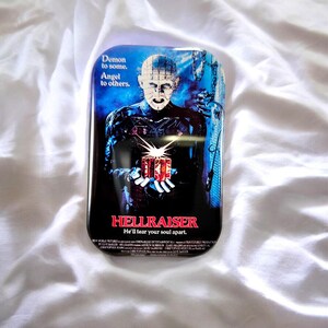 Könnte beinhalten: Eine rechteckige Metalldose mit dem Filmplakat von Hellraiser. Das Bild zeigt eine blasse Figur mit Nadeln im Kopf, die eine leuchtende Puzzlebox vor einem dunkelblauen Hintergrund hält. Der Text lautet: Demon to some. Angel to others. HELLRAISER He'll tear your soul apart.
