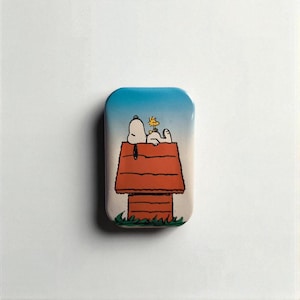 Könnte beinhalten: Eine rechteckige Anstecknadel mit Snoopy und Woodstock auf Snoopy's Hundehütte. Die Anstecknadel hat einen blauen Himmelshintergrund und ein rot-oranges Hundehäuschen. Das Gras ist grün.