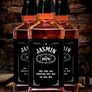 Könnte beinhalten: Drei Likörflaschen mit schwarzen Etiketten. Die Etiketten zeigen den Namen "Jasmin" und das Jahr "1976". Die Flaschen enthalten eine dunkelbernsteinfarbene Flüssigkeit und haben schwarze Verschlüsse. Der Text "Alles Liebe zum Geburtstag mein Hase Ich Liebe Dich" ist enthalten.