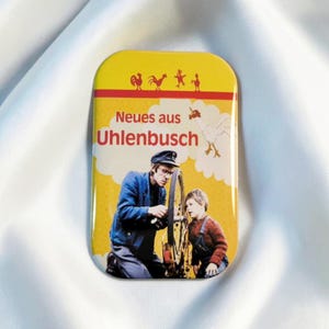 Könnte beinhalten: Ein rechteckiger Ansteckbutton mit gelbem Hintergrund und dem roten Schriftzug "Neues aus Uhlenbusch". Das Motiv zeigt eine Person in blauer Jacke und Mütze, die ein Fahrradrad neben einem Kind repariert, ergänzt durch kleine Hahn-Symbole.