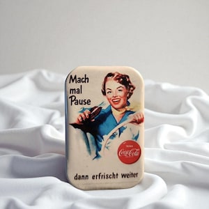 Könnte beinhalten: Eine rechteckige Metalldose mit einer Vintage-Illustration einer lächelnden Person, die eine Glasflasche Coca-Cola hält. Der Text lautet Mach mal Pause und dann erfrischt weiter, mit einem roten Coca-Cola-Logo in der unteren rechten Ecke.