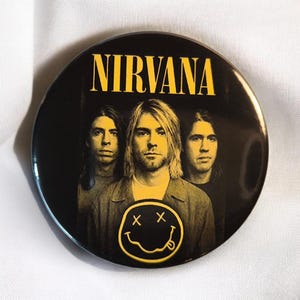 Könnte beinhalten: Ein runder Ansteckbutton mit einer schwarz-gelben Grafik der Band Nirvana. Das Design zeigt den gelben Schriftzug NIRVANA am oberen Rand, ein Porträt der drei Bandmitglieder in der Mitte und ein gelbes Smiley-Symbol im unteren Bereich.