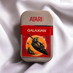 Könnte beinhalten: Eine rechteckige Metalldose mit dem Atari 2600-Logo oben und dem Spieltitel Galaxian in weißer Schrift auf rotem Grund. Die zentrale Grafik zeigt grüne Raumschiffe, die vor einer großen orangefarbenen Sonne im Weltraum fliegen.