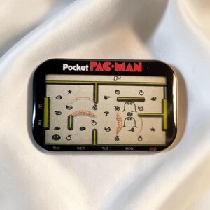 Könnte beinhalten: Ein rechteckiges, elektronisches Pocket PAC-MAN Handheld-Spiel mit schwarzem Rahmen und einem digitalen Bildschirm, der ein Labyrinth mit gelben Wänden, verschiedenen Fruchtsymbolen, zwei Geisterfiguren und die Zahl 04 oben mittig anzeigt.