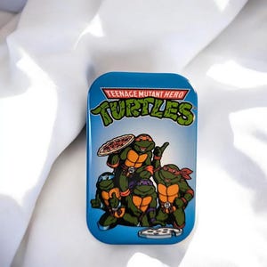 Könnte beinhalten: Eine rechteckige blaue Metalldose mit dem Teenage Mutant Hero Turtles Logo oben. Die Grafik zeigt vier grüne Schildkröten mit orangefarbenen Masken und Brustpanzern, eine hält eine Pizza, mit einem Pizzaschneider am unteren Rand.