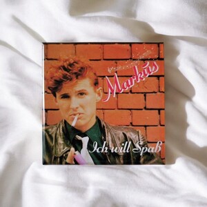 Könnte beinhalten: Ein quadratisches Musikalbum-Cover mit einer Person mit kurzem rötlichem Haar, die eine schwarze Lederjacke und ein dunkles Hemd trägt. Die Person hält eine Zigarette und ein rosa Feuerzeug. Der Hintergrund ist eine rote Backsteinmauer mit dem Text Markus und Ich will Spaß.