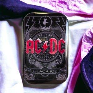 Könnte beinhalten: Eine rechteckige Metalldose mit einem komplexen schwarz-grauen Hintergrunddesign und dem roten AC/DC-Logo in der Mitte. Über dem Logo befindet sich ein kleines kreisförmiges Symbol eines Gitarristen, unter dem Logo steht der Text BLACK ICE in Weiß.