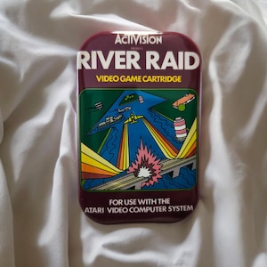 Könnte beinhalten: Eine rechteckige River Raid Videospielkassette für das Atari Video Computer System. Das Etikett zeigt das Activision-Logo, den Titel in weißer Schrift und eine bunte Grafik eines gelben Jets, der über einen Fluss mit Brücken und Schiffen fliegt.