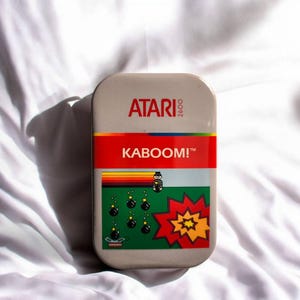 Könnte beinhalten: Eine rechteckige Metalldose mit dem Atari 2600-Logo oben und dem Schriftzug KABOOM! in Weiß auf rotem Grund. Darunter zeigt eine Grafik eine pixelige Figur, fallende Bomben, einen Regenbogenstreifen und eine große rot-gelbe Explosion.