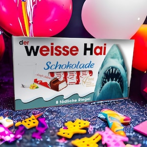 Könnte beinhalten: Eine Schachtel "der weisse Hai Schokolade" Schokoriegel mit einem Hai-Motiv. Die Schachtel enthält den Text "8 tödliche Riegel" und ein Bild eines Milchglases und eines Schokoriegels. Bunte Luftballons und Konfetti sind im Hintergrund.