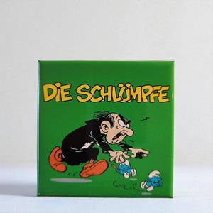 Könnte beinhalten: Eine quadratische grüne Fliese mit dem gelben Schriftzug Die Schlümpfe oben. Unter dem Text zeigt eine Comic-Illustration einen Mann in schwarzer Robe und orangefarbenen Schuhen, der nach zwei kleinen blauen Wesen auf grünem Grund greift.