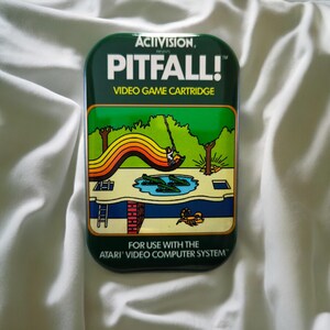 Könnte beinhalten: Eine rechteckige grüne Metalldose mit dem Artwork der Pitfall!-Videospielkassette. Die Grafik zeigt eine Figur, die an einer Liane über ein Krokodilbecken schwingt, mit dem Text ACTIVISION PRESENTS PITFALL! VIDEO GAME CARTRIDGE FOR USE WITH THE ATARI VIDEO COMPUTER SYSTEM.