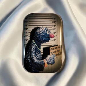Könnte beinhalten: Rechteckiges Tablett mit einer Parodie von Grover aus der Sesamstraße in einem Fahndungsfoto. Das blaue Monster hält ein Schild mit der Aufschrift "MONSTER, GRO, BK# 83-1124, Nov 24". Der Hintergrund hat eine Größentabelle.