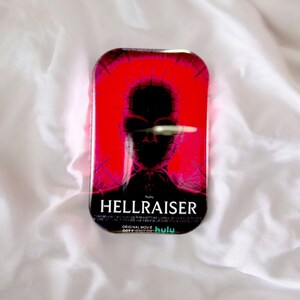 Könnte beinhalten: Eine rechteckige Metalldose mit leuchtend rotem Hintergrund und der dunklen Silhouette einer Figur mit Nadeln im Kopf. Der Text HELLRAISER ist in Weiß gedruckt, ergänzt durch weiteren Text wie Hulu und Original Movie.