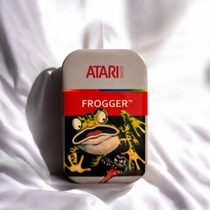 Könnte beinhalten: Eine rechteckige Metalldose mit dem Artwork des Atari 2600 Videospiels Frogger. Das Design umfasst das rote Atari 2600 Logo, ein rotes horizontales Band mit dem Text FROGGER und eine Grafik eines grünen Frosches über einem kleinen Alligator.