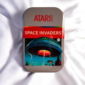 Könnte beinhalten: Eine rechteckige Metalldose mit dem Atari 2600 Logo in Rot oben. Ein rotes Querband zeigt den Text SPACE INVADERS in Weiß. Darunter zeigt eine Grafik ein großes blaues UFO, das über einer Landschaft mit kleineren fliegenden Untertassen schwebt.