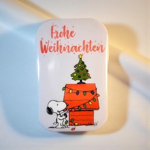 Könnte beinhalten: Eine rechteckige weiße Metalldose mit einer Abbildung von Snoopy, der eine rote Hundehütte mit bunten Weihnachtslichtern schmückt. Ein grüner Weihnachtsbaum steht auf dem Dach und der Text Frohe Weihnachten ist in Rot darüber gedruckt.