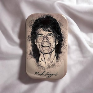 Könnte beinhalten: Eine rechteckige Metalldose mit einem schwarz-weißen Porträt von Mick Jagger im Skizzenstil. Das Kunstwerk verwendet dunkle, ausdrucksstarke Linien, um die Gesichtszüge und Haare einzufangen. Der Name Mick Jagger steht in Schreibschrift am unteren Rand.