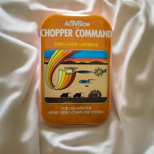 Könnte beinhalten: Eine orangefarbene, rechteckige Activision Chopper Command Videospielkassette für das Atari Video Computer System. Das Etikett zeigt eine bunte Grafik mit einem Hubschrauber, einem Regenbogen und Bodenfahrzeugen sowie weißem Text auf orangefarbenem Grund.