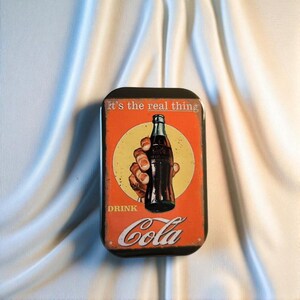 Könnte beinhalten: Eine rechteckige Metalldose mit abgerundeten Ecken und orangefarbenem Hintergrund. Das Design zeigt eine Hand, die eine dunkle Glasflasche in einem gelben Kreis hält, mit dem in Weiß gedruckten Text It's the real thing und DRINK Cola.