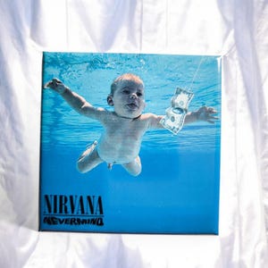 Könnte beinhalten: Ein quadratisches Vinyl-Schallplattencover mit dem ikonischen Nevermind-Albumcover von Nirvana. Das Bild zeigt ein Baby, das unter Wasser auf einen Dollarschein an einem Haken zuschwimmt, vor einem hellblauen Hintergrund mit Band- und Albumtitel.