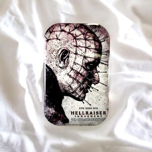 Könnte beinhalten: Eine rechteckige Metalldose mit einem Grafikdruck der Figur Pinhead aus dem Film Hellraiser: Judgement. Das Bild zeigt das Profil eines blassen, strukturierten Kopfes mit zahlreichen Nadeln in der Haut. Der Text lautet Evil Seeks Evil, Hellraiser und Judgement.