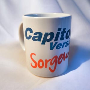 Könnte beinhalten: Weiße Keramiktasse mit blauem und rotem Text. Der Text lautet "Capitol Version" und "Sorgeu".
