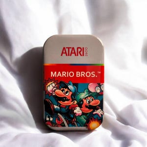 Könnte beinhalten: Eine rechteckige Metalldose mit dem Atari 2600 Logo oben und dem Schriftzug Mario Bros. in Weiß auf rotem Grund. Der untere Teil zeigt eine farbenfrohe grafische Illustration von Mario und Luigi in Action-Posen mit verschiedenen Gegnern.