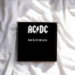 Könnte beinhalten: Ein quadratisches schwarzes Albumcover mit dem weißen AC/DC-Logo oben und einem Blitz in der Mitte. Unter dem Logo ist der Text BACK IN BLACK in einer klaren, weißen Serifenschrift in Großbuchstaben auf dem einfarbigen schwarzen Hintergrund gedruckt.