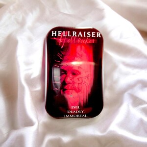 Könnte beinhalten: Eine rechteckige Metalldose mit einer dunkelroten und schwarzen Grafik einer Figur mit Nadeln im Kopf. Der Text HELLRAISER Hellseeker ist oben aufgedruckt, und die Wörter EVIL. DEADLY. IMMORTAL. sind unten auf der Dose zu sehen.