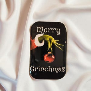 Könnte beinhalten: Ein rechteckiger schwarzer Magnet mit einer grünen, pelzigen Hand mit einem weiß abgesetzten roten Ärmelbündchen, die eine glänzende rote Weihnachtskugel hält. Der Text "merry" steht in Weiß darüber und "GRINCHMAS" steht in Weiß darunter.