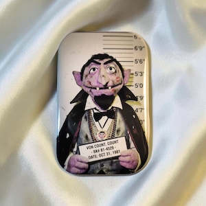 Könnte beinhalten: Ein rechteckiger Magnet mit einem Fahndungsfoto von Graf Zahl aus der Sesamstraße. Graf Zahl trägt einen Smoking, einen Umhang und Schmuck und hält ein Schild mit der Aufschrift "VON COUNT, COUNT - BK# 81-4529 - DATE: OCT 31, 1981". Der Hintergrund hat eine Größentabelle.