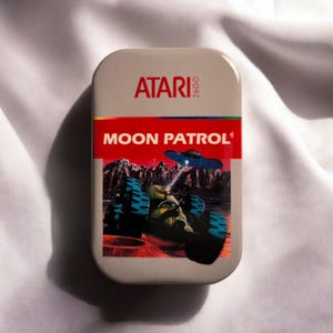 Könnte beinhalten: Eine rechteckige beige Metalldose mit dem Atari 2600 Logo oben und dem Spieltitel Moon Patrol in weißer Schrift auf rotem Grund. Die Grafik zeigt einen grünen Mondrover mit blauen Reifen auf einer roten Oberfläche unter einem dunklen Himmel.