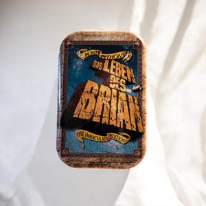 Könnte beinhalten: Eine rechteckige Metalldose mit dem Filmtitel Monty Python's Das Leben des Brian und dem Untertitel The Immaculate Edition. Das Design zeigt große, texturierte goldene Schriftzüge auf blauem Grund mit einem steinartigen Rahmen.