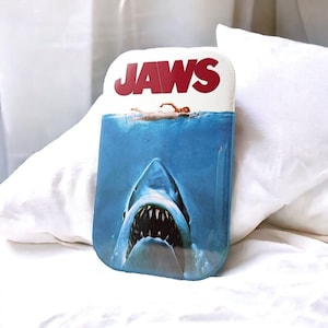 Könnte beinhalten: Eine rechteckige Metalldose mit dem Filmplakat von Jaws. Das Design zeigt einen großen Hai, der nach oben in Richtung einer auf der Wasseroberfläche schwimmenden Person schwimmt, wobei das rote Jaws-Logo oben auf dem Behälter abgedruckt ist.