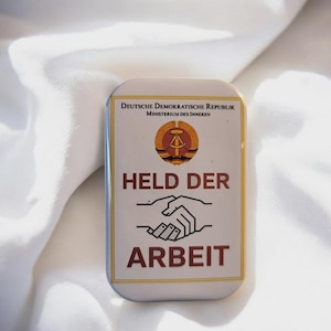 Könnte beinhalten: Rechteckige Anstecknadel mit abgerundeten Ecken, mit dem Text "HELD DER ARBEIT" und einem Emblem. Die Anstecknadel hat einen weißen Hintergrund mit goldenem Rand und die Worte "DEUTSCHE DEMOKRATISCHE REPUBLIK" oben.