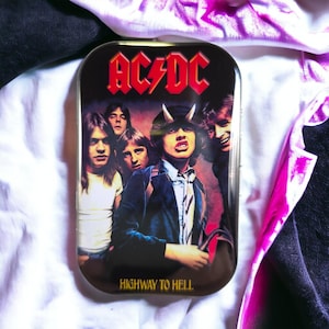 Könnte beinhalten: Eine rechteckige Metalldose mit dem AC/DC-Logo in Rot oben und dem Schriftzug Highway to Hell unten. Die Oberfläche zeigt eine Grafik der Bandmitglieder, wobei die zentrale Figur kleine Hörner auf dem Kopf trägt.