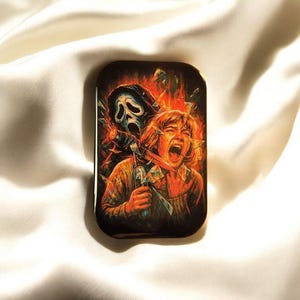 Könnte beinhalten: Eine rechteckige Metalldose mit einer lebendigen, detaillierten Illustration einer maskierten Figur mit weißem, geisterhaftem Gesicht, die hinter einer schreienden Person steht. Die Szene ist vor einem feurigen orange-roten Hintergrund mit Glassplittern dargestellt.