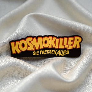 Könnte beinhalten: Ein schwarzer Emaille-Pin mit dem Wort KOSMOKILLER in fetten gelben Buchstaben mit roter Umrandung. Unter dem Haupttext steht der Schriftzug SIE FRESSEN ALLES in einer kleineren gelben Schrift. Der Pin hat einen schwarzen Hintergrund und Glanz.